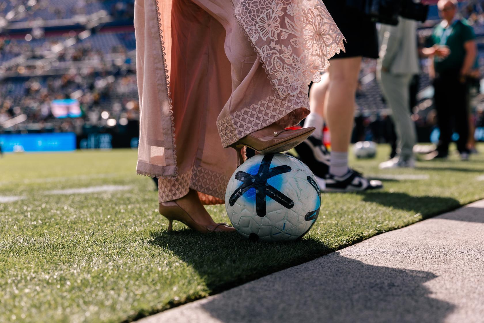 A person wearing heels is seen standing at the side of a football pitch with one heel standing on a football