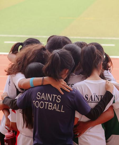 A team is seen hugging with their backs to the camera, one team member has the word Saints Football written on the back of their jersey