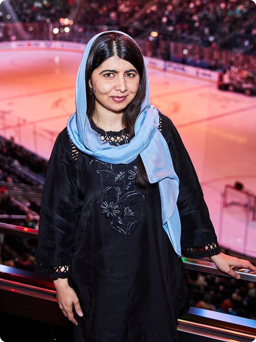 Malala Yousafzai is photographed in the stands of a ice rink stadium with the stadium visible in the background