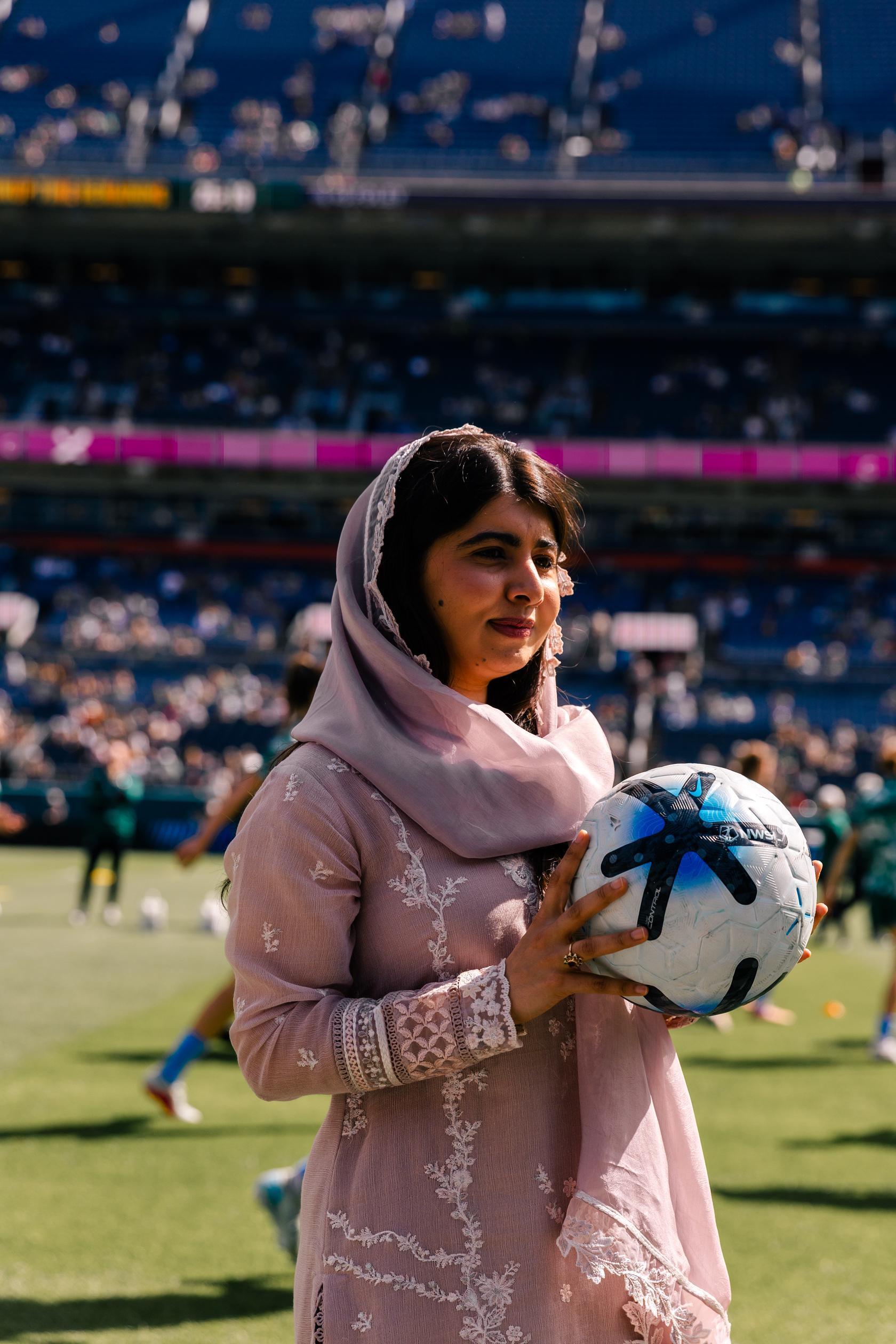 Malala Yousafzai is seen holding a football standing on a pitch the stadium and football memebers are seen in the background which has been slightly blurred