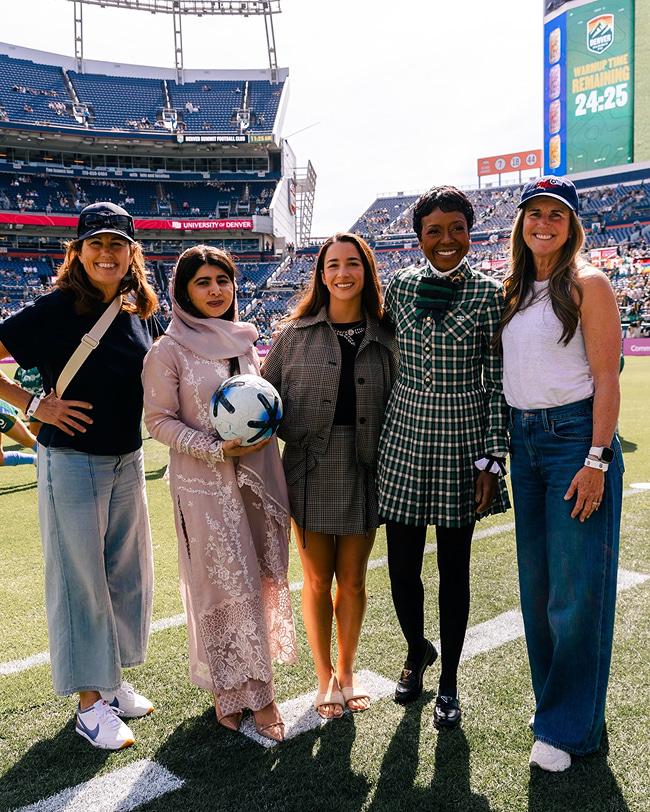 Malala is seen standing among a group holding a football, all are facing the camera smiling and standing on the pitch of a stadium with the stadium seats visible in the background.