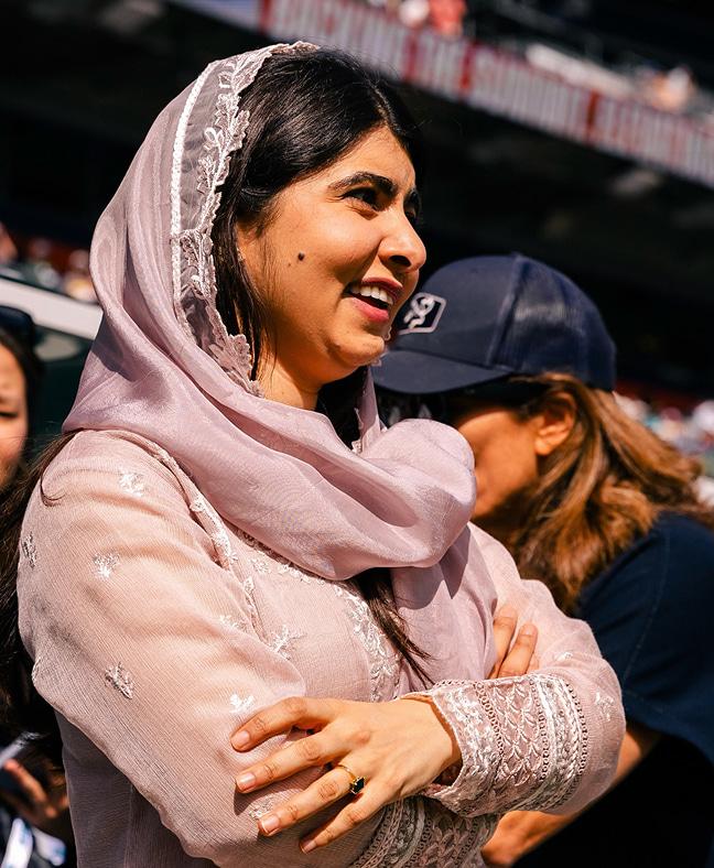 Malala is seen standing among a crowd in a stadium looking out and smailing.