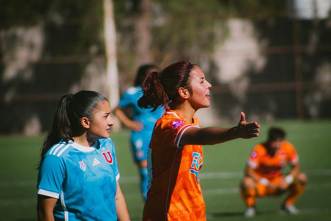 Two players are seen standing on the pitch, one player has a blue jersey and the other has an orange jersey. The player in orange is motioning with their right hand extended to the side.