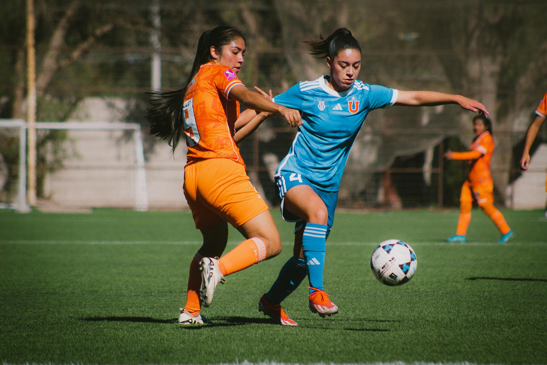 Two football players are seen mid game. One player has an orange jersey and the other play has a blue. The orange player can be seen mid tackle with some team members visible in a blurred background.