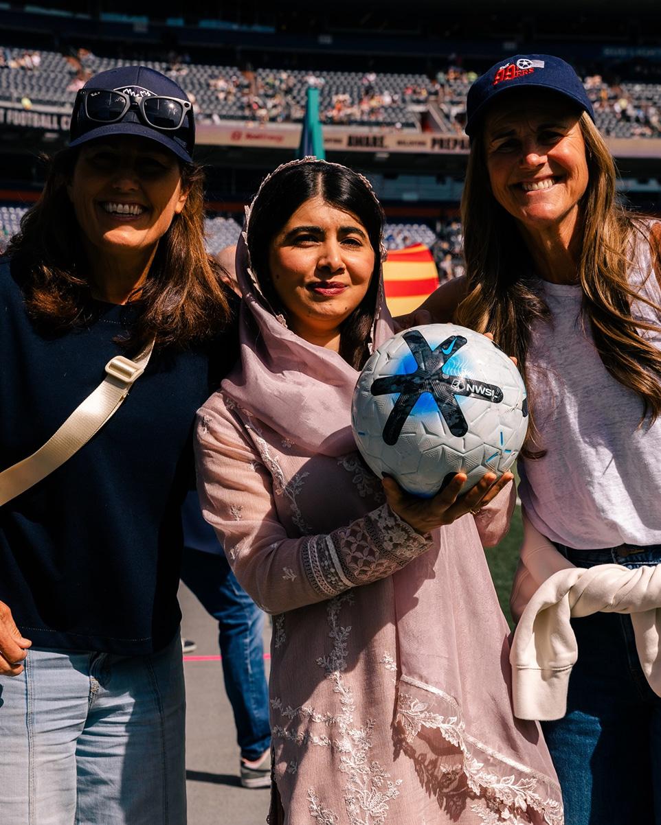 Malala is seen standing in a group of smiling people holding a football, there is a person either side of her and both are wearing caps.